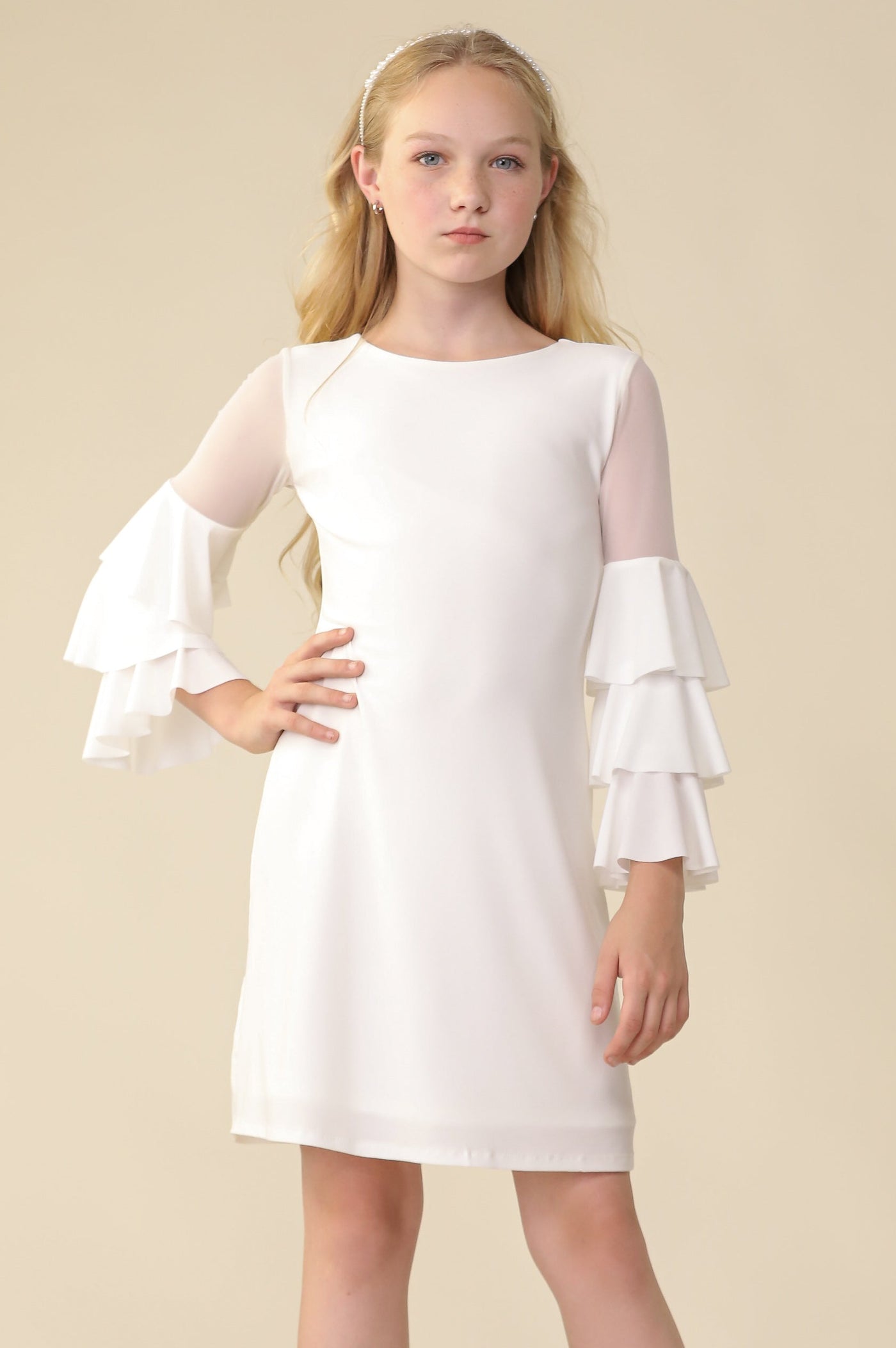 A young girl with blonde hair stands against a solid beige studio background wearing a white knee-length dress with sheer long sleeves and tiered ruffle cuffs, paired with cream lace-up ankle boots.