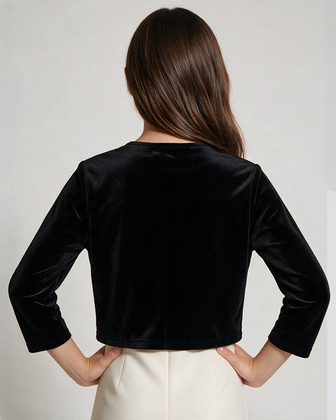 A woman viewed from the back wearing a black velvet long-sleeve cropped top and cream-colored pants, set against a plain light gray studio background.