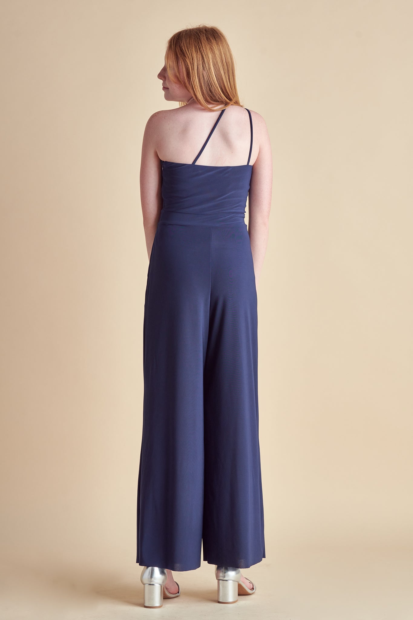 Blonde girl in a one shoulder, navy wide leg jumpsuit with silver heel.