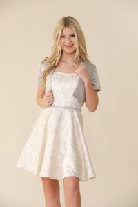 A young blonde haired girl in the fit and flare silver and ivory dress that hits above the knee with a silver bolero jacket..
