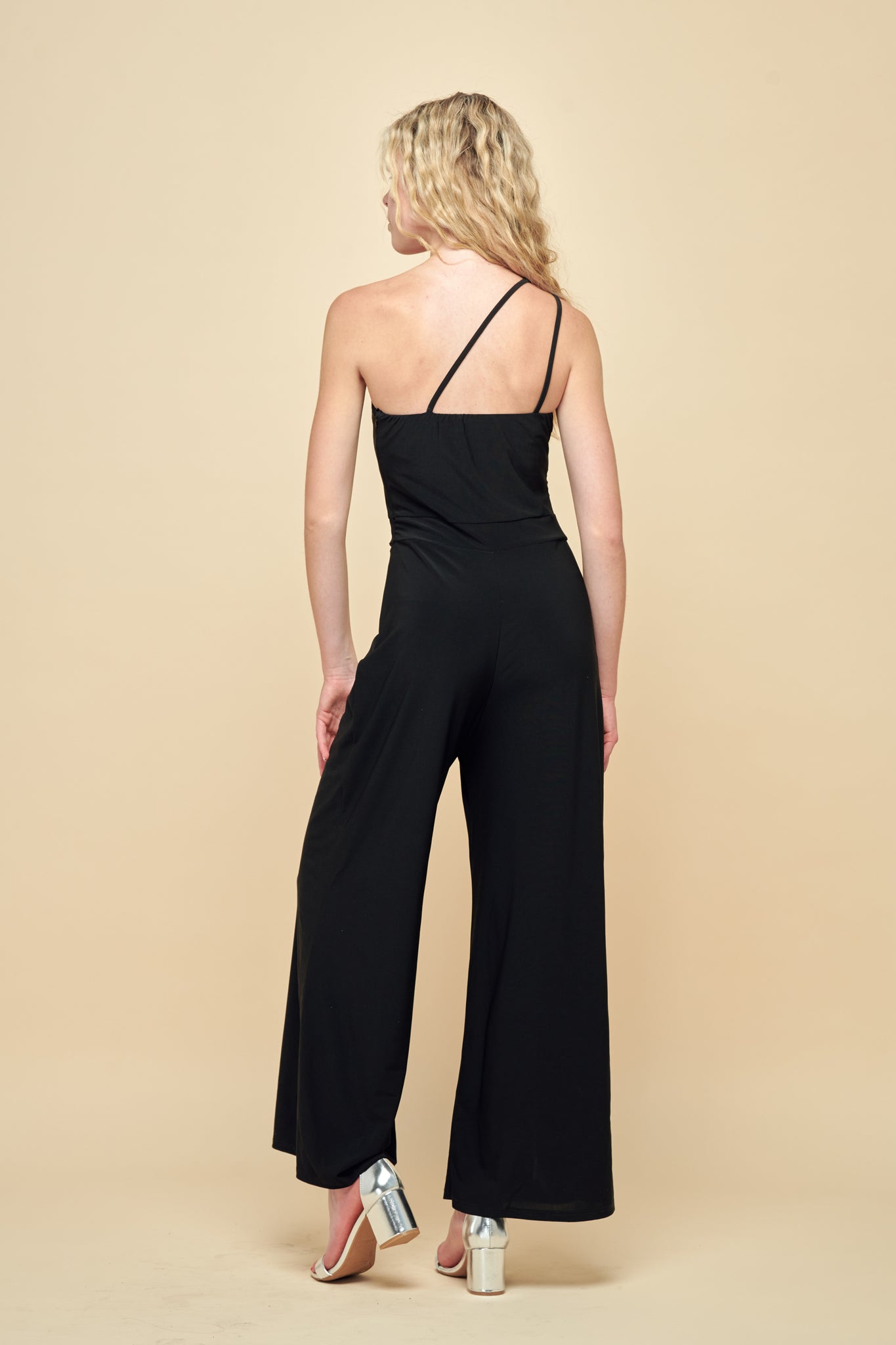 Blonde girl in a one shoulder, wide leg jumpsuit with silver heel.